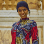 Fakhriya stands smiling at the camera wearing a head scarf and brightly patterned dress