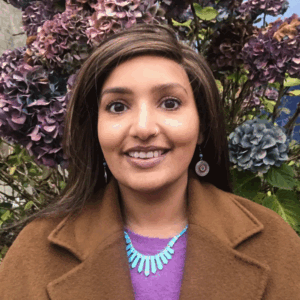 Humayra stands smiling at the camera with her hair down and wears a purple top, with brown coat